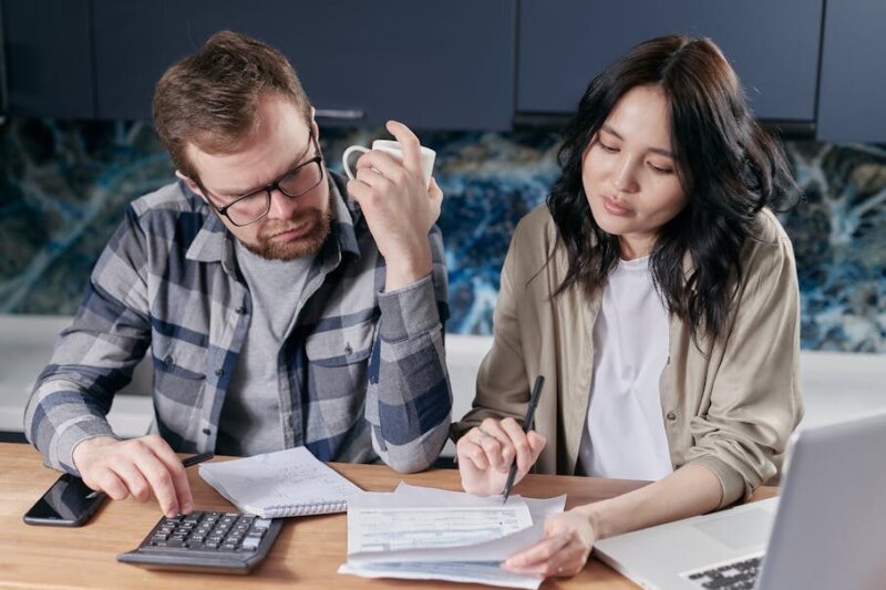 A couple reviewing household bills and budget using a calculator and laptop at their kitchen table.